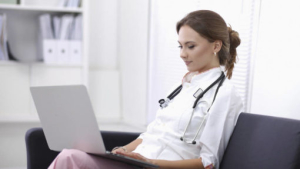 Female doctor with a stethoscope around her neck sitting in an office chair and working on a laptop.