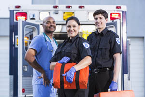 Three emergency medical professionals stand in front of an open ambulance, smiling and holding medical equipment.