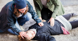 Two people assisting a woman who has collapsed on the ground, one checking her pulse and the other supporting her head, suggesting a medical emergency or first aid situation.