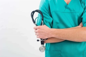 Medical professional in green scrubs holding a stethoscope, standing with arms crossed against a plain white background.