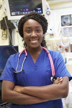 Smiling nurse in blue scrubs with a stethoscope around her neck stands with arms crossed in a hospital or medical environment.