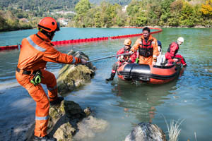 Rescue workers in orange suits pull a boat with teammates to shore on a river, surrounded by trees and floating barriers.