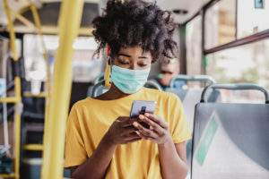 Woman wearing a face mask and yellow shirt using her smartphone while sitting on a bus.