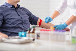 Healthcare worker preparing to draw blood from a patient's arm, with medical supplies and test tubes visible on the table.