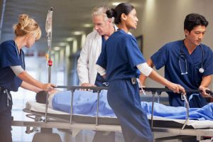 Medical team in scrubs and a doctor rush a patient on a gurney through a hospital corridor, with IV attached, appearing to respond to an emergency.