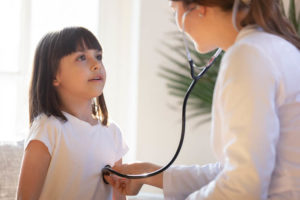 Female doctor using stethoscope to listen to young girl's chest during a medical checkup in a brightly lit room.