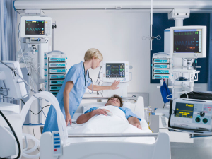 Nurse monitoring a patient in a hospital bed surrounded by medical equipment and vital sign monitors in an intensive care unit.