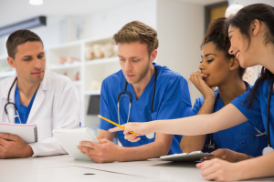 Group of medical students and a doctor in scrubs discussing notes and collaborating around a table in a classroom or hospital setting.