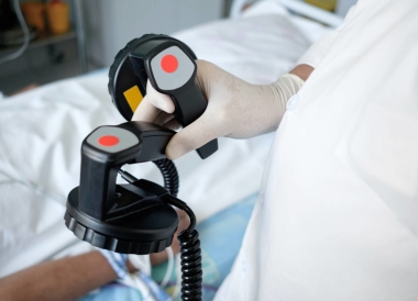 Medical professional wearing gloves holding defibrillator paddles, preparing to use them on a patient lying in a hospital bed.