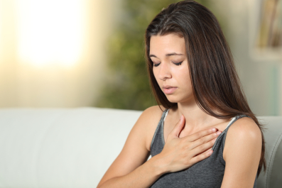 Young woman sitting on a couch with her hand on her chest, eyes closed, appearing to experience chest discomfort or pain.